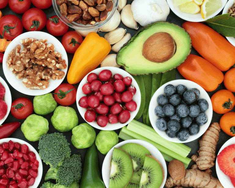 An assortment of fresh fruit, vegetables, nuts, and spices arranged closely together, including avocado, blueberries, tomatoes, kiwi, ginger, garlic, broccoli, walnuts, and lemon slices.