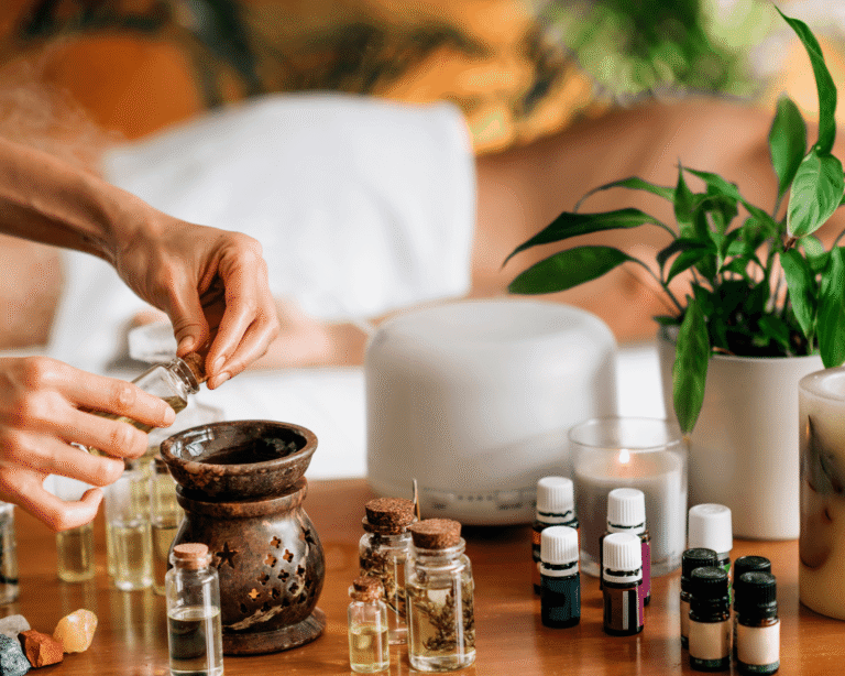 Hands prepare an essential oil burner on a wooden table with bottles of oils, a lit candle, crystals, and a potted plant. A person lies face down on a massage table in the blurred background.