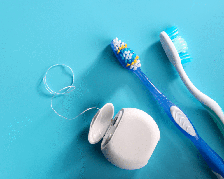 Two toothbrushes and an open container of dental floss are arranged on a light blue surface. Some dental floss is pulled out of the container and lies loosely on the surface.