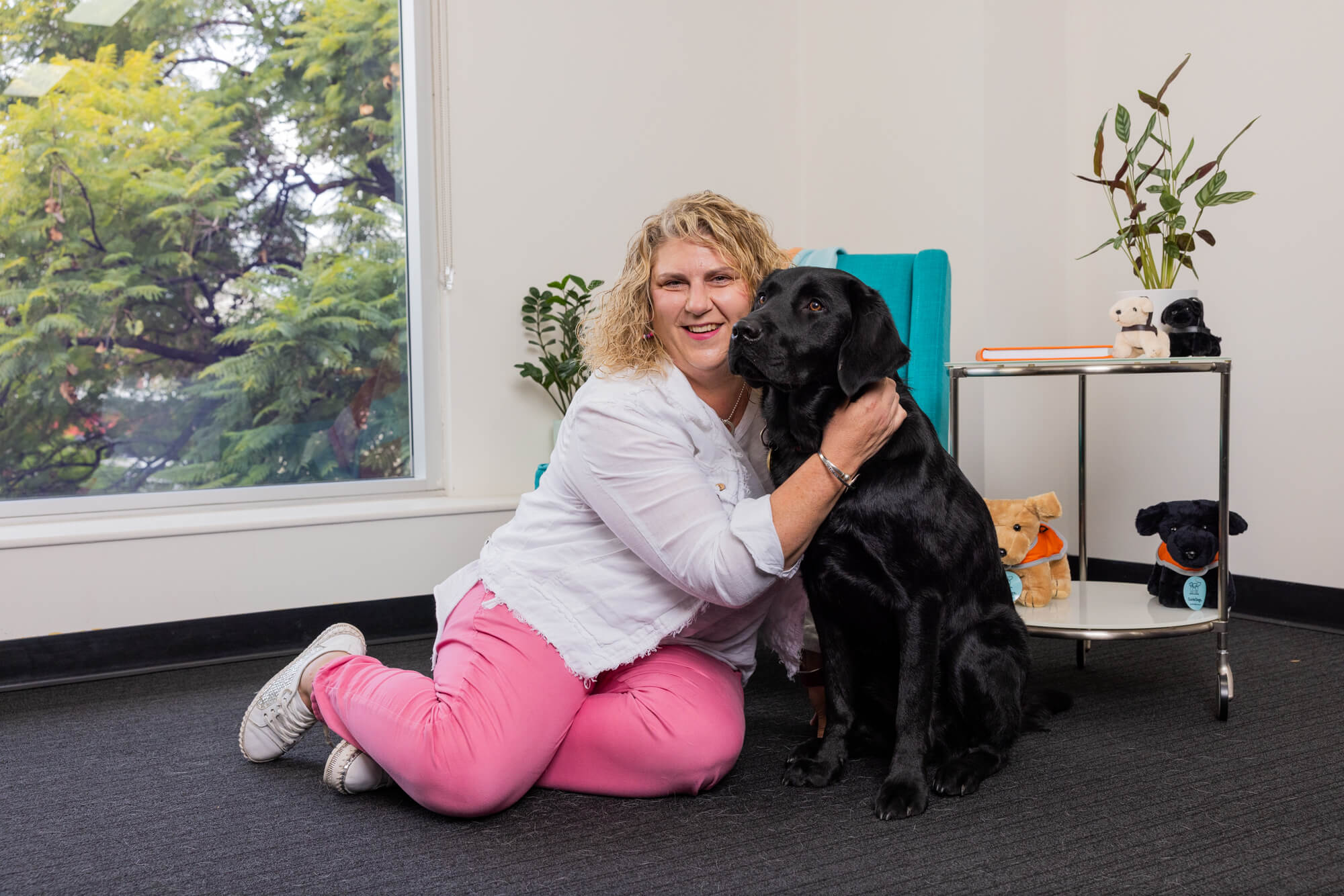 A woman with curly blonde hair wearing a white shirt and pink trousers sits on the floor indoors, smiling and hugging a black Labrador guide dog. There is a small shelf with stuffed dogs, books, and a plant nearby, as well as a window with trees outside—a warm scene that speaks to the lasting impact of leaving a gift in your will to support guide dogs.