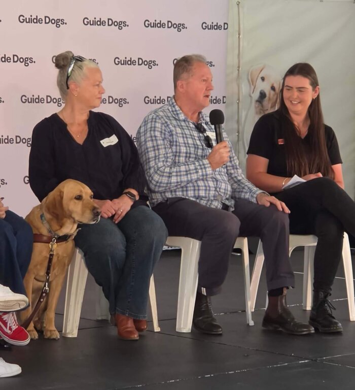 Two women and a man sit on chairs on a stage in front of a Guide Dogs branded backdrop, with a yellow Labrador guide dog sitting next to one of the women.
