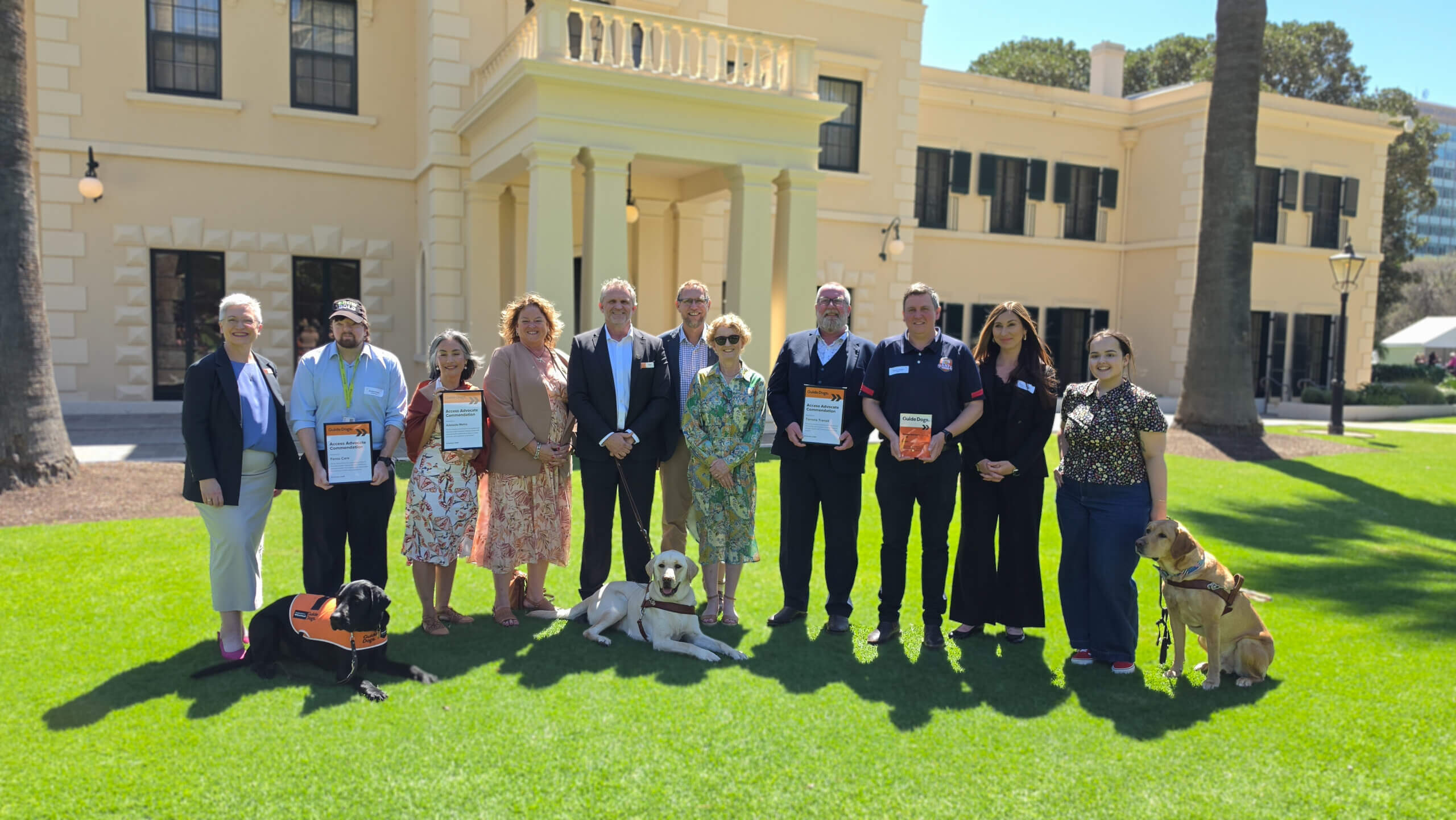 A group of people standing on a lawn in front of a beige building, some holding certificates and books, with two assistance dogs resting on the grass.