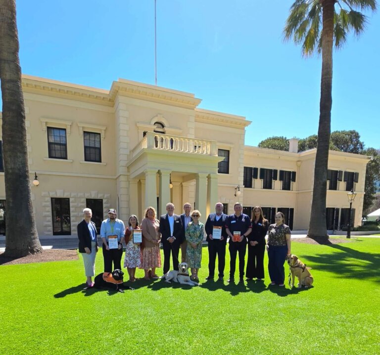 A group of eleven people and two dogs stand in a line on a lawn in front of a large, pale building.
