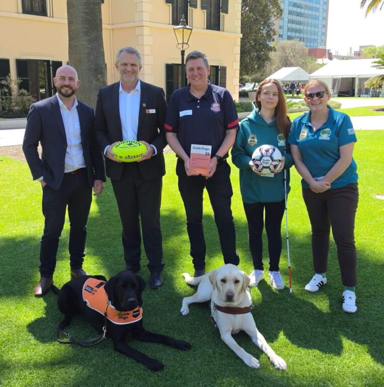 Three men and two women stand on a lawn with two guide dogs resting at their feet; the people are holding two sports balls and a Guide Dogs Australia information booklet.
