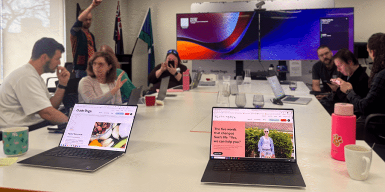 A group of people sit around a long meeting table with open laptops, facing a large wall-mounted screen displaying a colourful graphic.