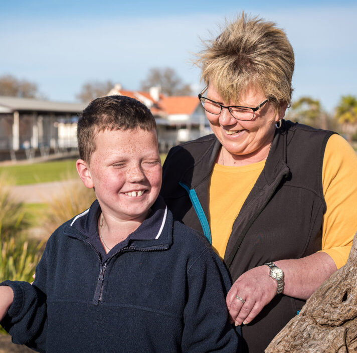 A young boy and his mum are smiling and standing in a park.