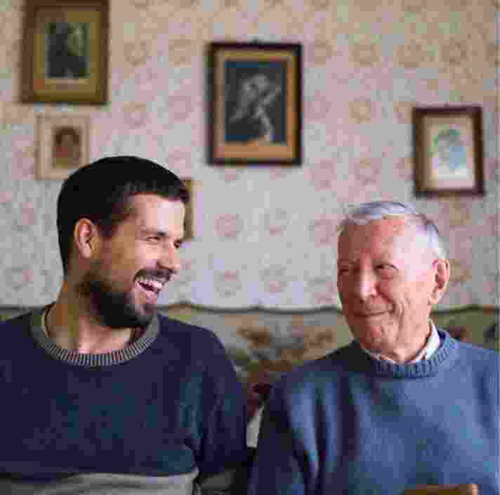 Two people, one adult & one older person, sitting on a couch looking at each other laughing.