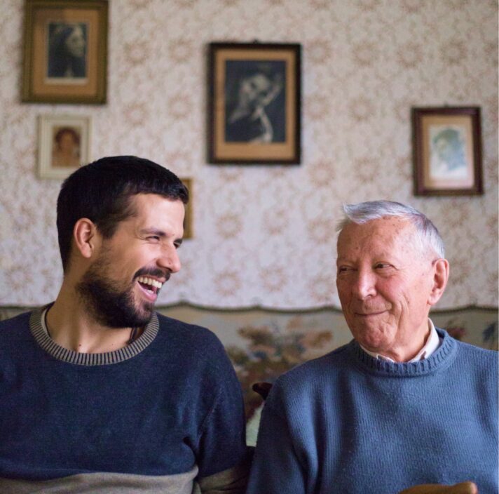 Two people, one adult & one older person, sitting on a couch looking at each other laughing.