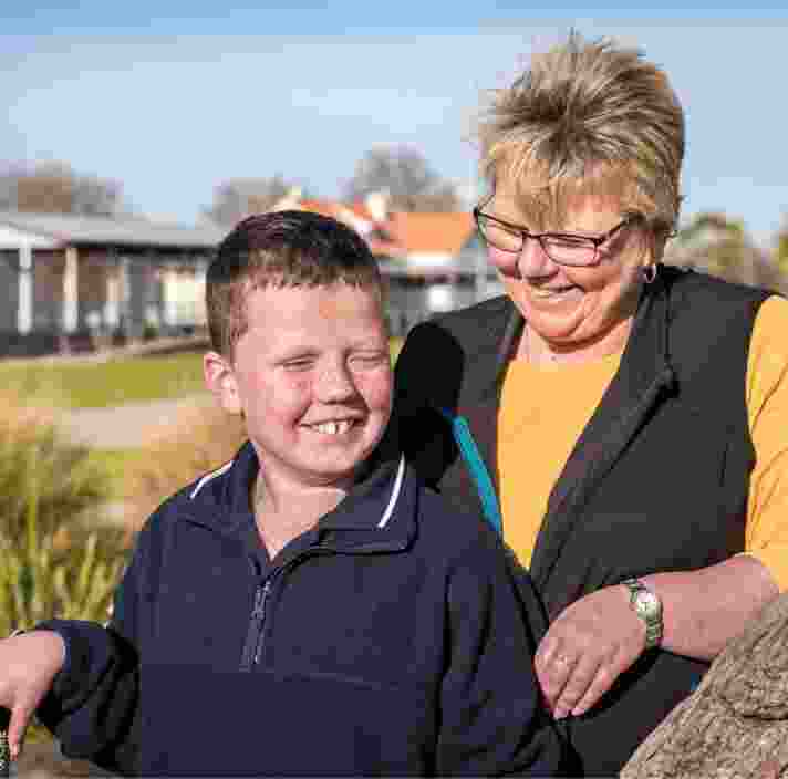A young boy standing outside with his mother. The mother is close behind her son and they are both smiling, The mother is looking at her son.