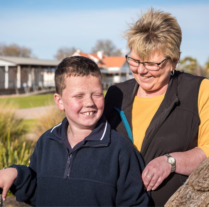 A young boy standing outside with his mother. The mother is close behind her son and they are both smiling, The mother is looking at her son.