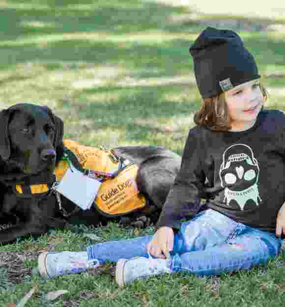 A young child sitting outside on some grass next to an Autism Assistance Dog. The dog is a black labrador and is wearing an Autism Assistance Dog jacket.