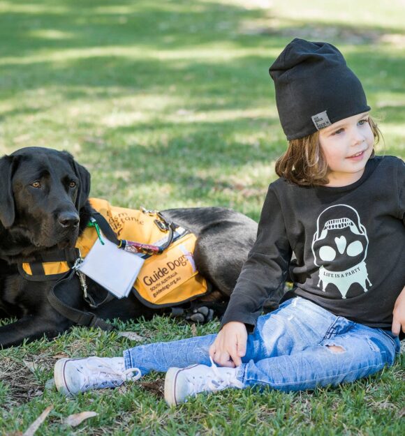 A young child sitting outside on some grass next to an Autism Assistance Dog. The dog is a black labrador and is wearing an Autism Assistance Dog jacket.