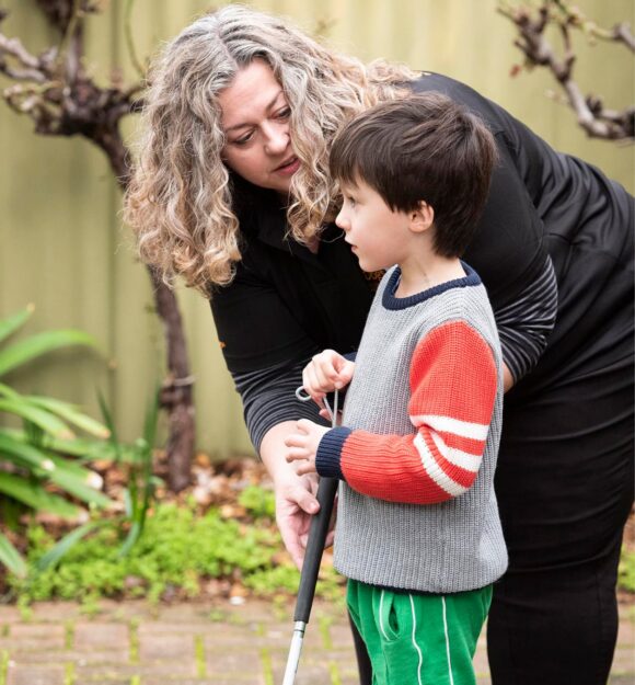 A young child using their white cane outside while an adult assists them.