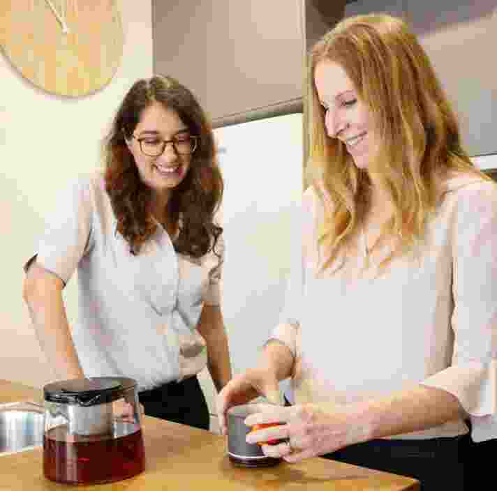 Two people inside in the kitchen. One is making tea while the other is watching on.