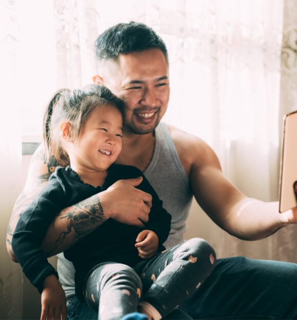 A young child sitting on their father's lap while they both smile while looking at an Ipad. The father has one had around his child and is holding the Ipad with the other hand.