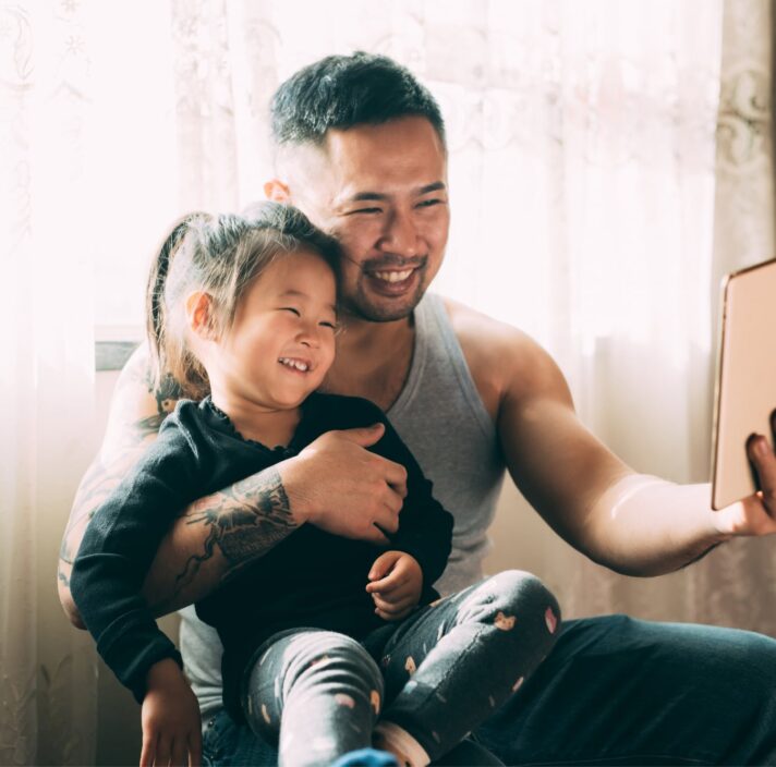A young child sitting on their father's lap while they both smile while looking at an Ipad. The father has one had around his child and is holding the Ipad with the other hand.