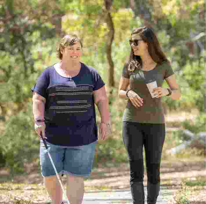 Two people walking outside. One is using a white cane and the other is holding a drink. They are both smiling.