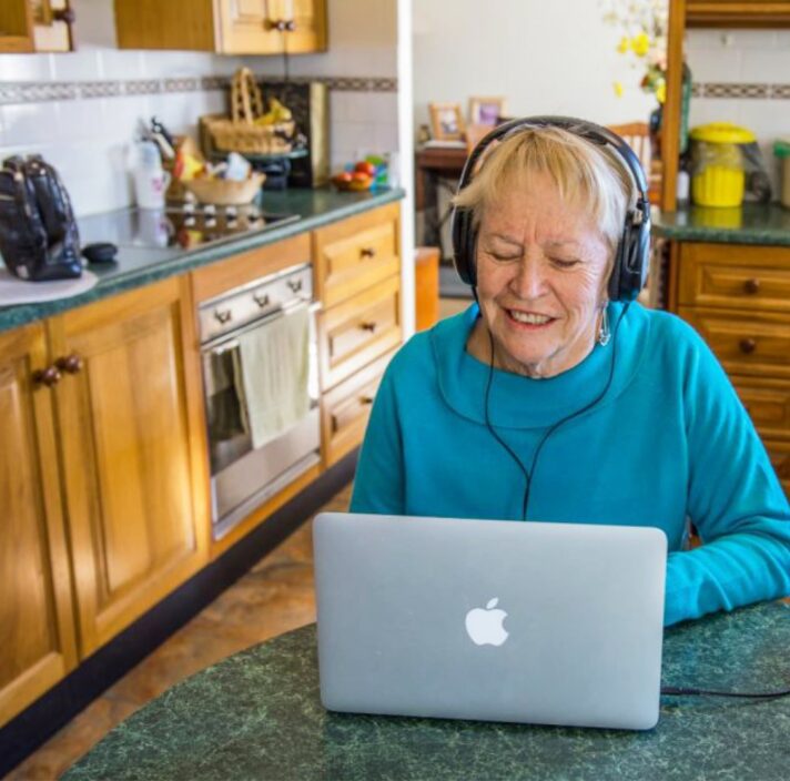 An older adult wearing headphones whilst on the laptop. The person is seated at a kitchen table.