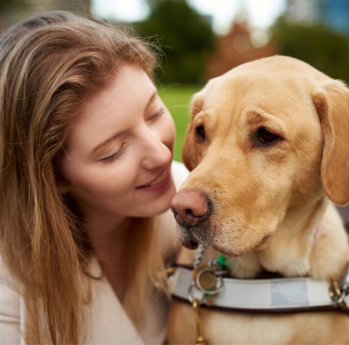 Close up of a person with their Guide Dog, who is caramel in colour and in harness. The person is smiling at the dog.