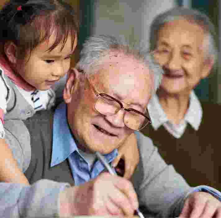 An older man writing while a young girl puts her arms around his shoulders, hugging him. The man is smiling and there is an older woman in the background who is not in focus but is also smiling.
