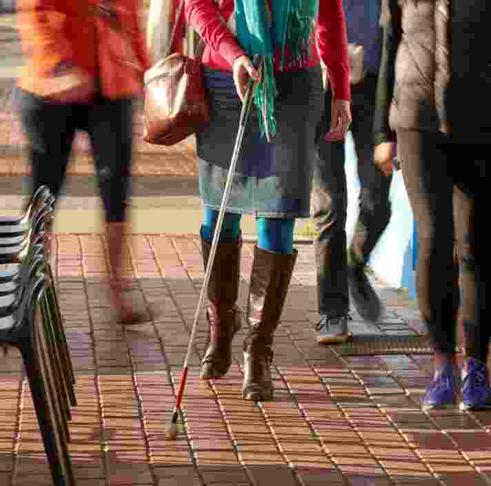 A person walking down the street using their cane whilst there are other people walking next to them. The image is from the waist down