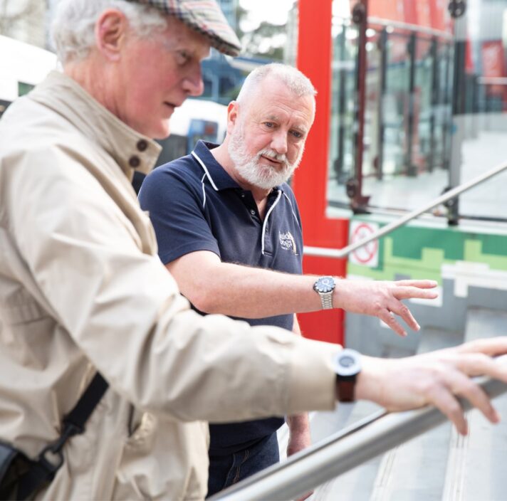 A Guide Dogs staff member assisting an an older adult with navigating a handrail.