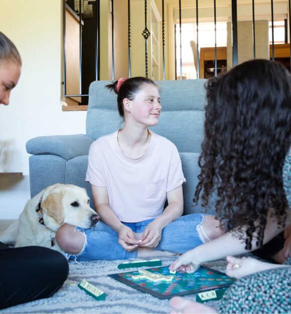 A group of four young adults sitting inside, in a circle. There is a yellow labrador dog who is resting its head on one of the person's legs.