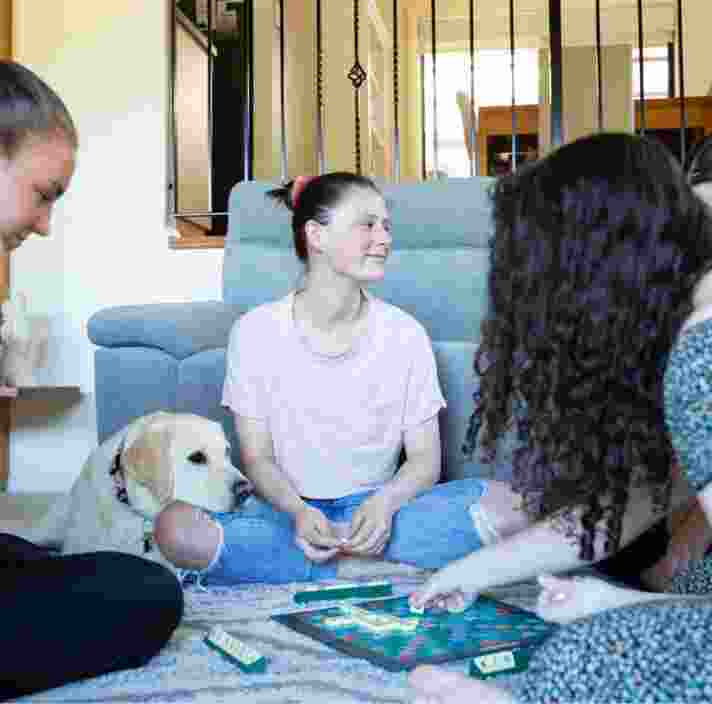 A group of four young adults sitting inside, in a circle. There is a yellow labrador dog who is resting its head on one of the person's legs.