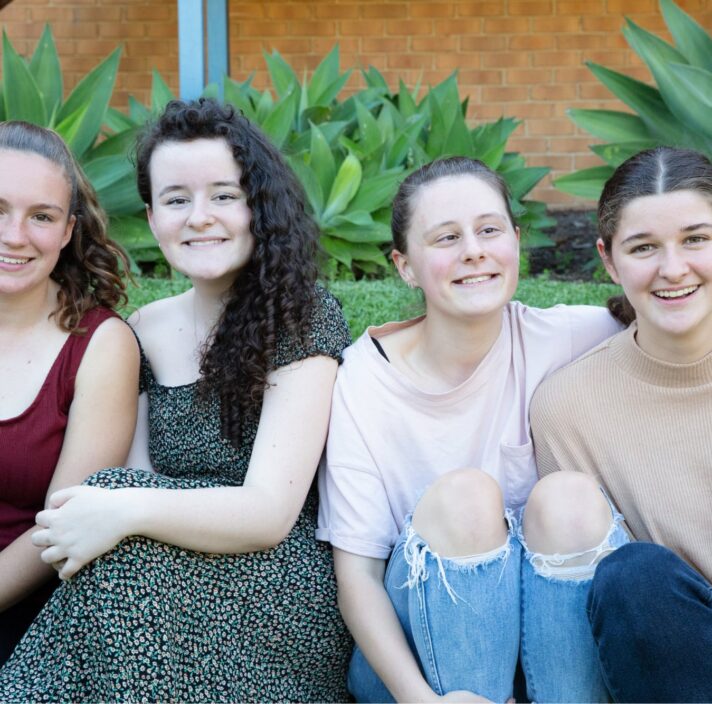 A group of four young adults sitting outside on the grass all in a row. Everyone is looking at the camera smiling.