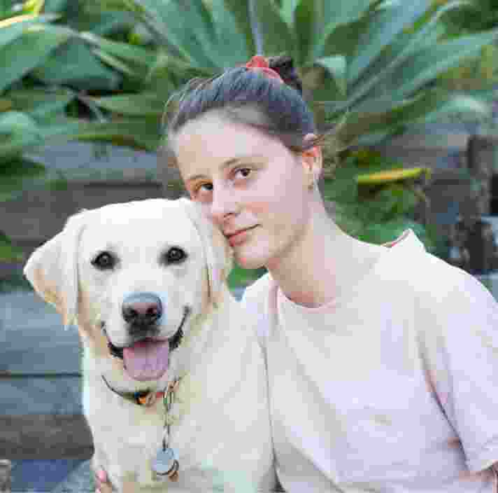 A person looking straight at the camera with their arm around a yellow labrador dog. They are seated at the bottom of a set of stairs.