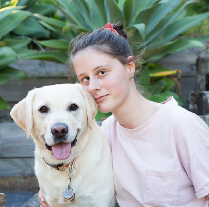 A person looking straight at the camera with their arm around a yellow labrador dog. They are seated at the bottom of a set of stairs.