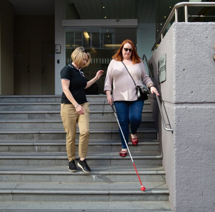 A person assisting a person using a white cane down a set of stairs.