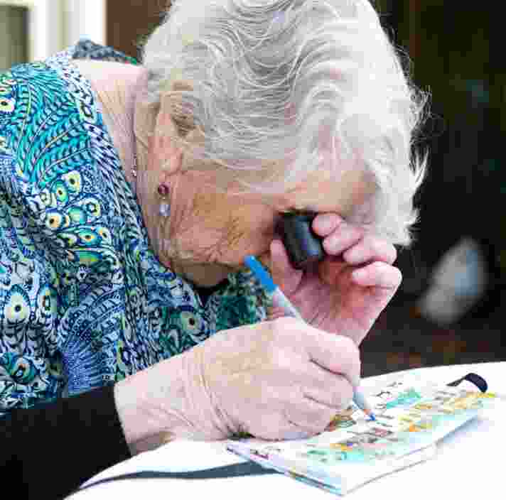 An older adult using a magnifier whilst painting, The person is painting onto a canvas.