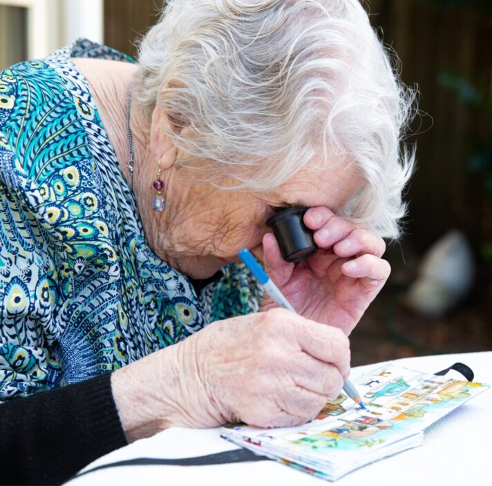 An older adult using a magnifier whilst painting, The person is painting onto a canvas.