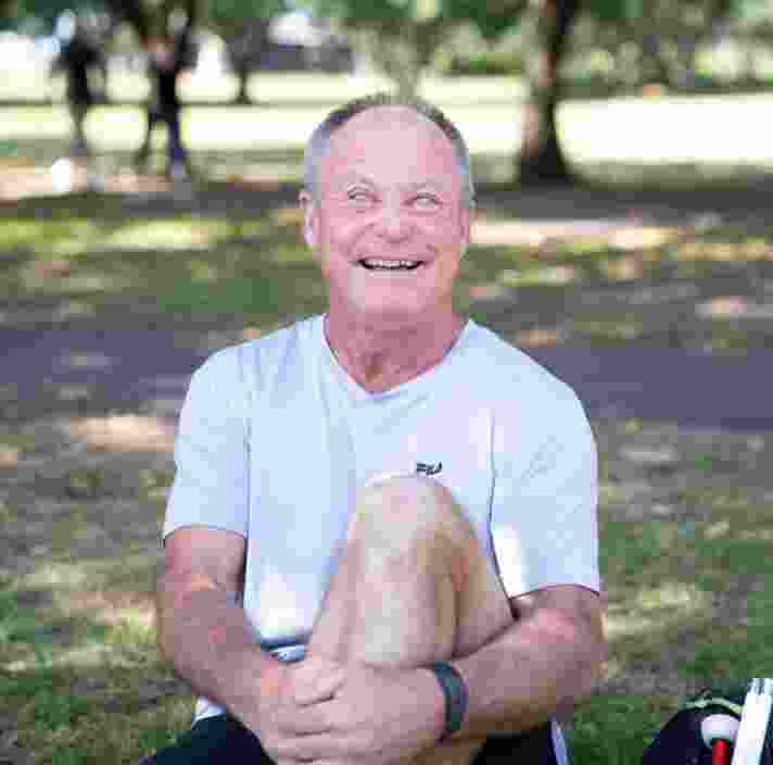 A person sitting outside on the grass with a smile on their face looking at the camera. They have one leg bent with their arm wrapped around their knee and a white cane next to them.