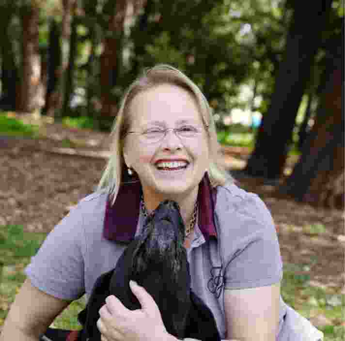A person and their black labrador Guide Dog outside in the park. The dog is looking up at the person and the person is smiling at the camera.
