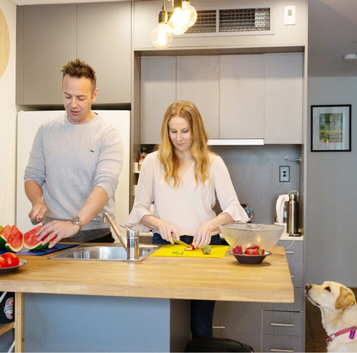 A couple preparing fresh food in their kitchen. Next to the kitchen table is a yellow labrador dog sitting on its back legs looking up at the food.