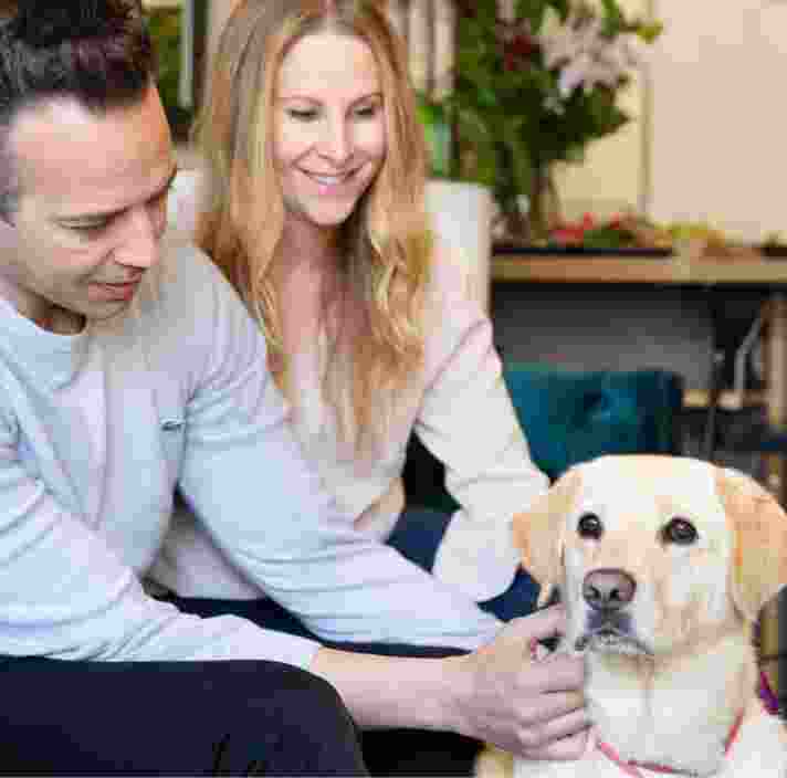 Two people sitting on their couch patting their yellow labrador dog.