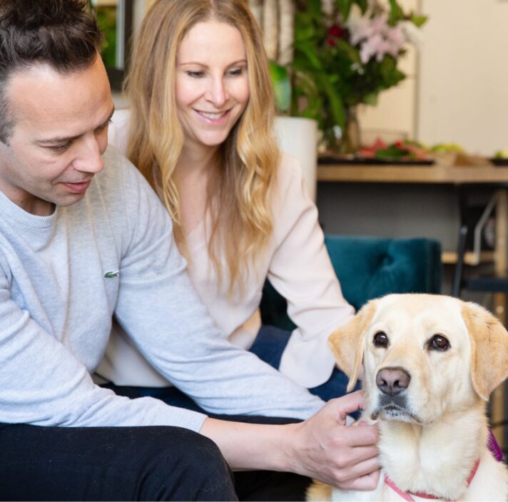 Two people sitting on their couch patting their yellow labrador dog.