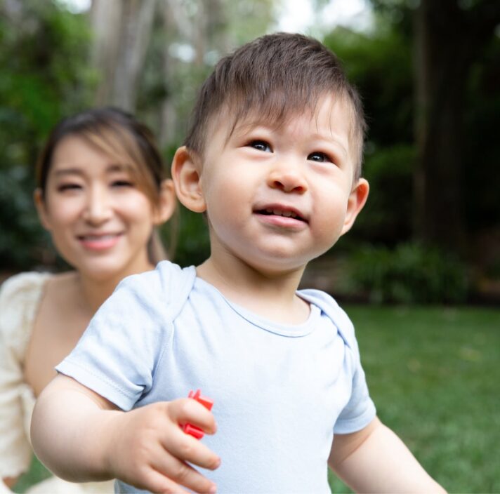 A young child playing outside with the mother watching in the background.