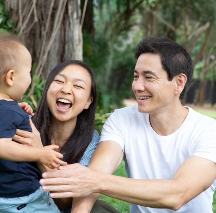 A couple sitting outside watching their baby walk towards them. The mother and father have smiles on their faces.