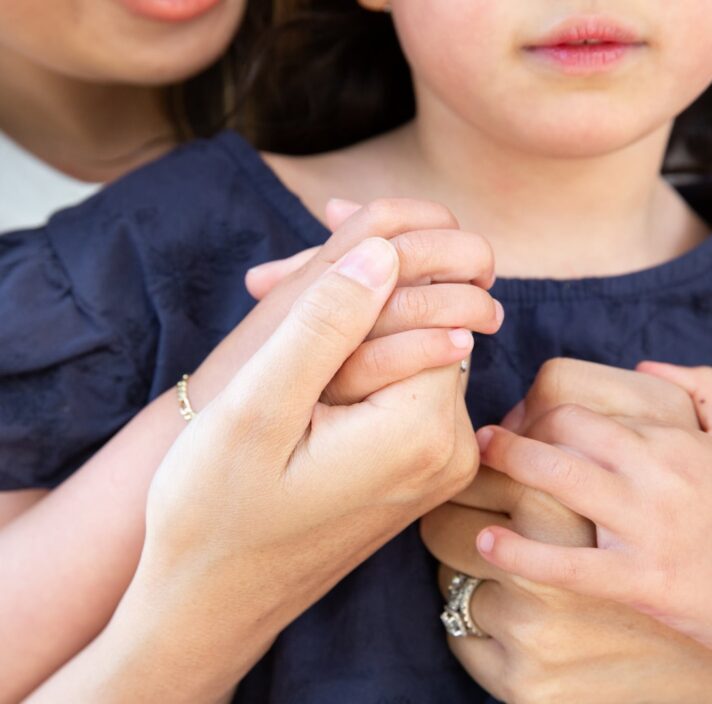 A close up of a mother cuddling a young child from behind, their hands are embraced.