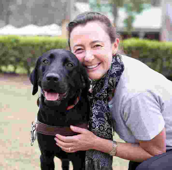 A person hugging a black labrador Guide Dog in harness. The person and dog are looking at the camera and the person is smiling,