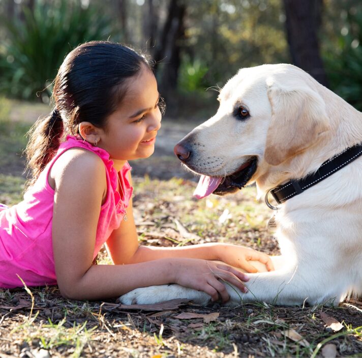 A young child laying down looking into the face of a yellow labrador dog with a smile on their face, They are both outside and are sitting facing each other.