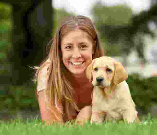 A person laying on some grass with an eight week old yellow labrador puppy. The person is smiling and looking at the camera. The puppy is also looking at the camera.