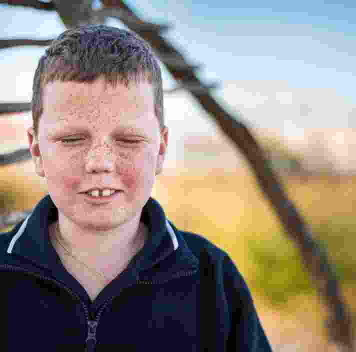 A young child standing outside looking at the camera. The child has their eyes closed.
