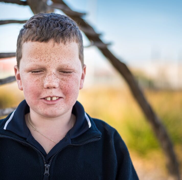 A young child standing outside looking at the camera. The child has their eyes closed.