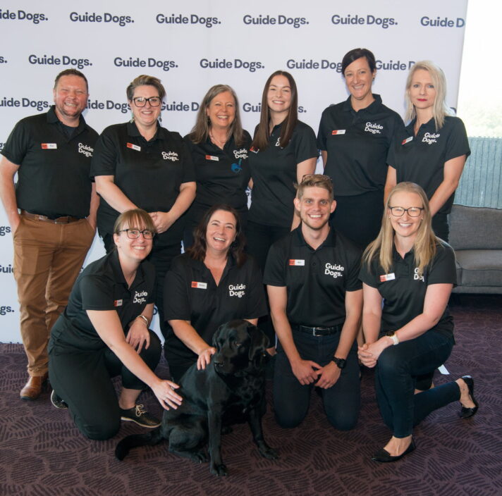 A group of Guide Dogs SA/NT staff members smiling at the camera. At the centre of the group is a black Labrador, Ambassadog Esky.