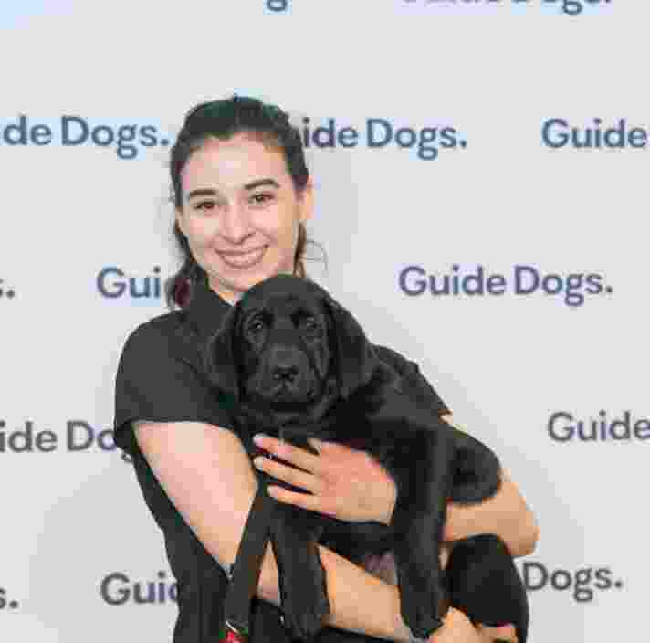A person in black clothing smiling at the camera and holding a small black Labrador puppy in their arms.