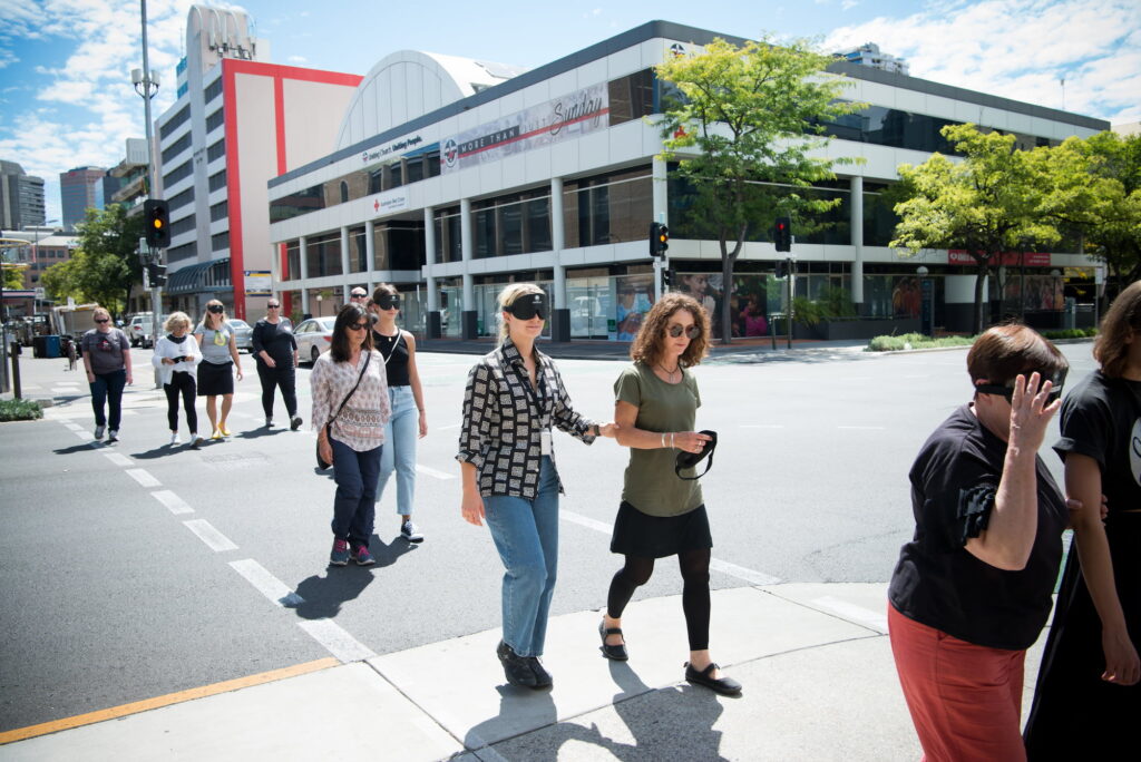 People walking in pairs cross an Adelaide street at a pedestrian crossing. One person in each pair is wearing a blindfold and holding on to the other.